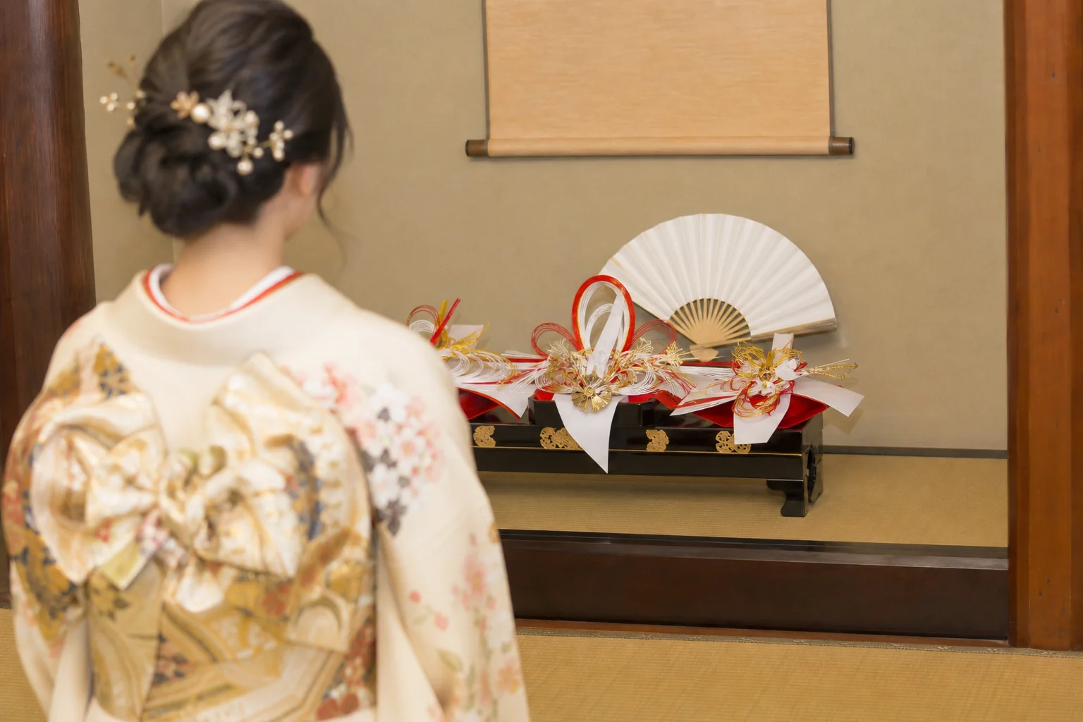 振袖の袖口と帯の後ろ姿が、和室の床の間の結納品の前に見える写真